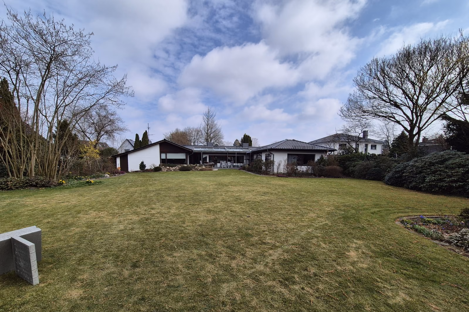 Große, eben angelegte Rasenfläche hinter einem Bungalow, umlaufend von Beeten und Gehölzen gefasst